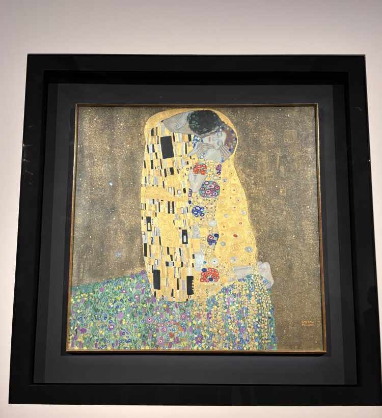 The Kiss by Gustav Klimt; painting of two people in an embrace wrapped around in gold draping